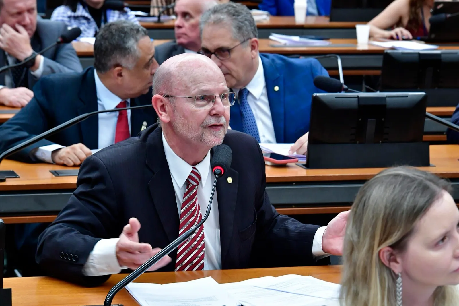 Parlamentar de terno escuro fala ao microfone durante reunião da Comissão Mista de Orçamento, com colegas sentados ao fundo.