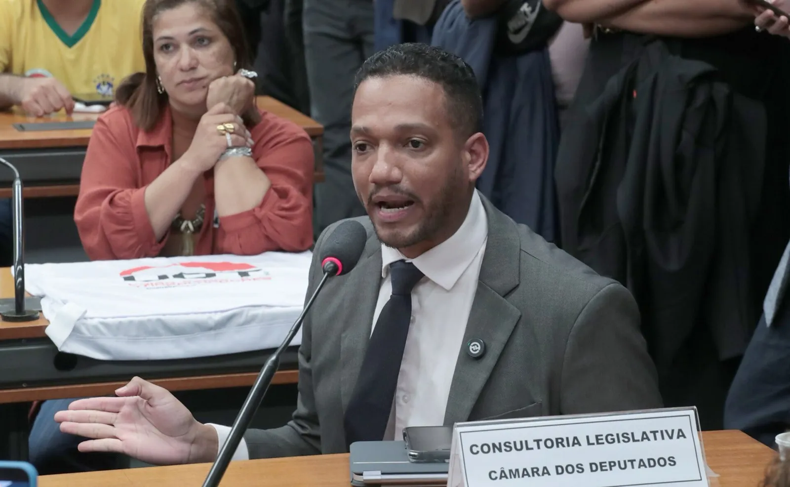Homem de terno cinza e gravata escura fala ao microfone numa sala de comissão da Câmara, diante de uma placa da Consultoria Legislativa; ao fundo, uma mulher e outras pessoas assistem.