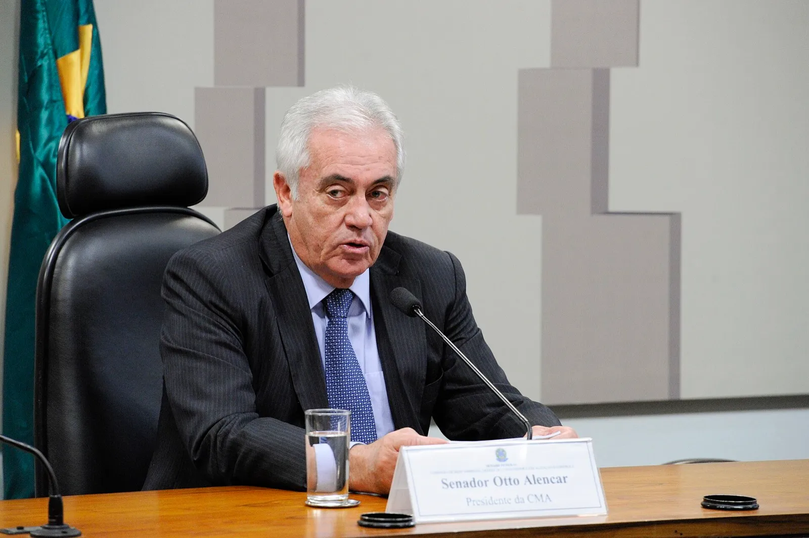 Senador Otto Alencar, de terno escuro, fala ao microfone na mesa da Comissão de Meio Ambiente do Senado, com a bandeira do Brasil ao fundo.