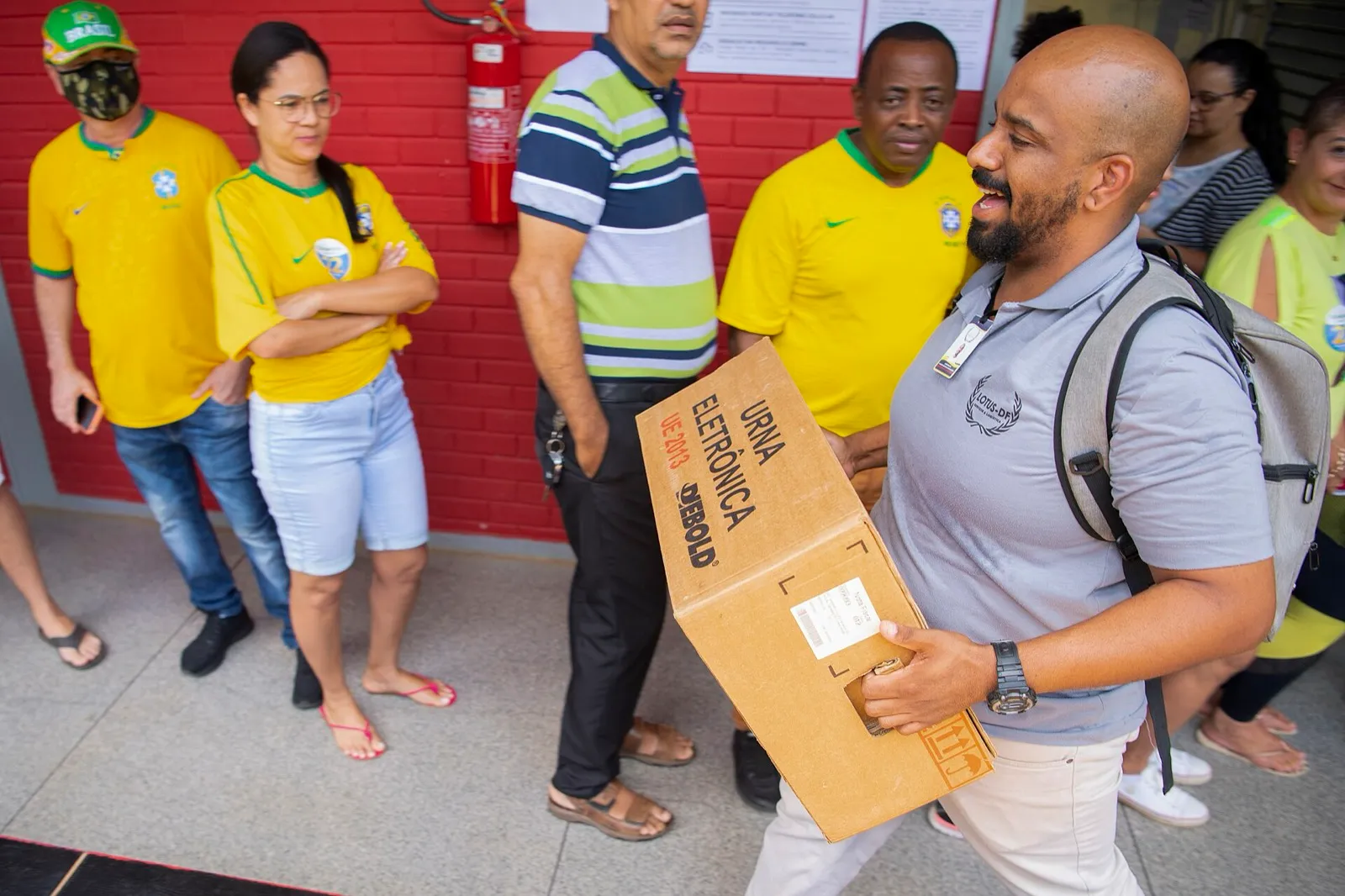 Homem carrega caixa de papelão com a inscrição 'urna eletrônica' em frente a uma escola, com eleitores de camisas amarelas em fila ao fundo.
