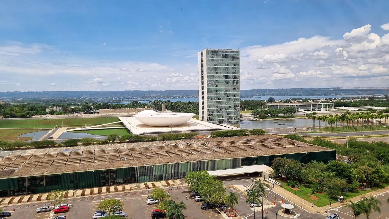Vista aérea do Palácio do Congresso Nacional, em Brasília, com a cúpula branca em primeiro plano, a torre de vidro ao centro e céu azul com nuvens.