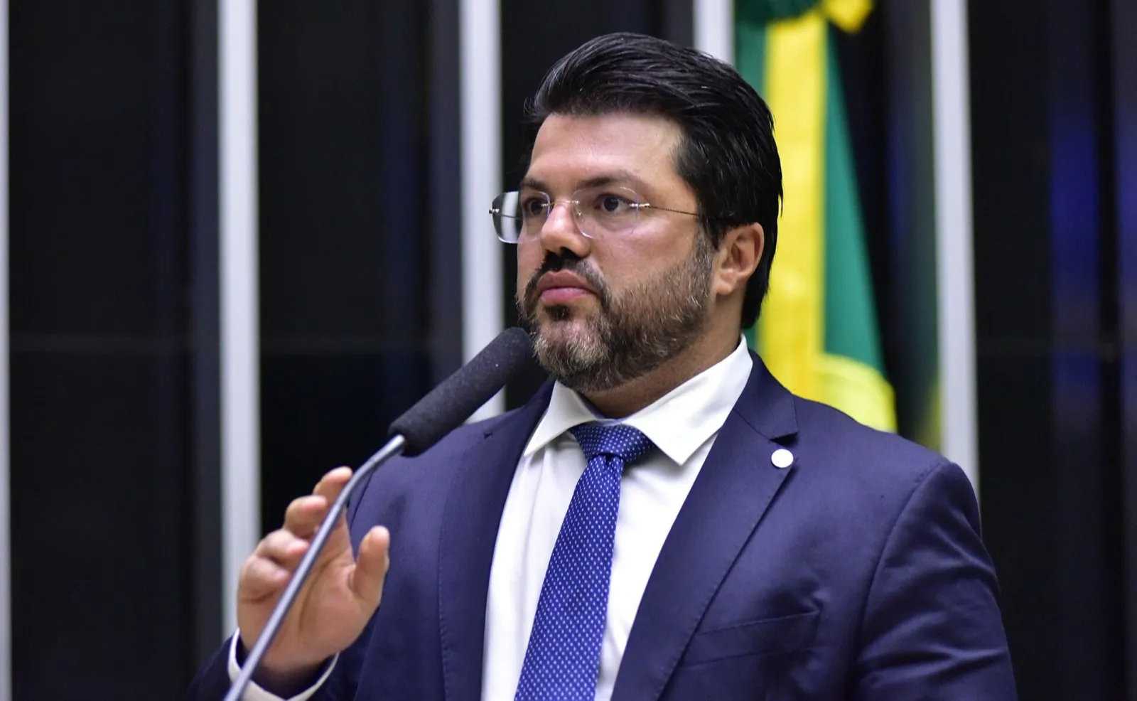 Deputado de terno azul fala ao microfone no plenário da Câmara, com a bandeira do Brasil ao fundo.
