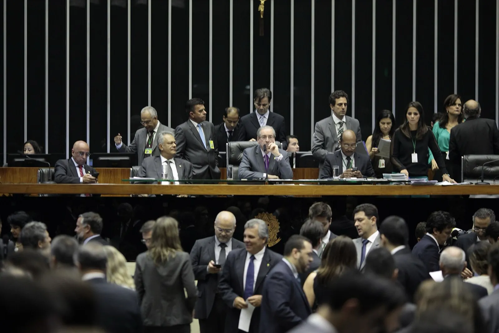Plenário da Câmara dos Deputados, com deputados e funcionários nas mesas e na área de circulação, sob um painel escuro com colunas metálicas.
