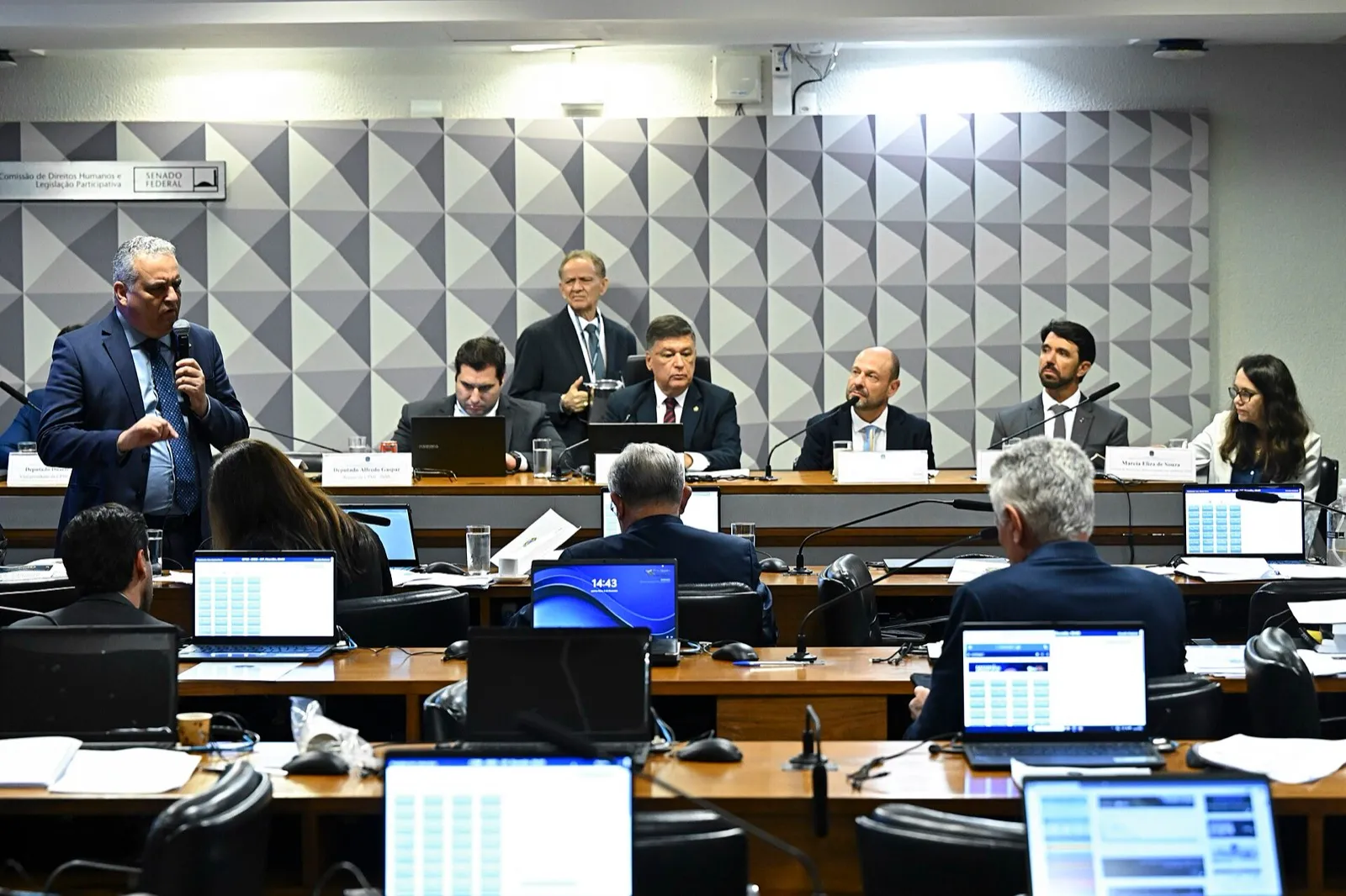 Sessão da CPMI do INSS no Senado: integrantes sentados à mesa diretora, um parlamentar de pé falando ao microfone e outros em bancadas com notebooks.