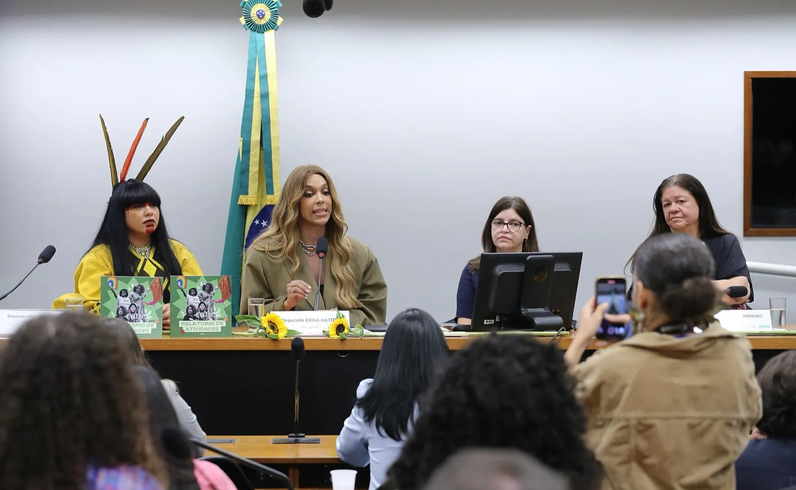 Mesa de uma reunião de comissão da Câmara dos Deputados, com a bandeira do Brasil ao fundo, mulheres à mesa e pessoas na plateia.