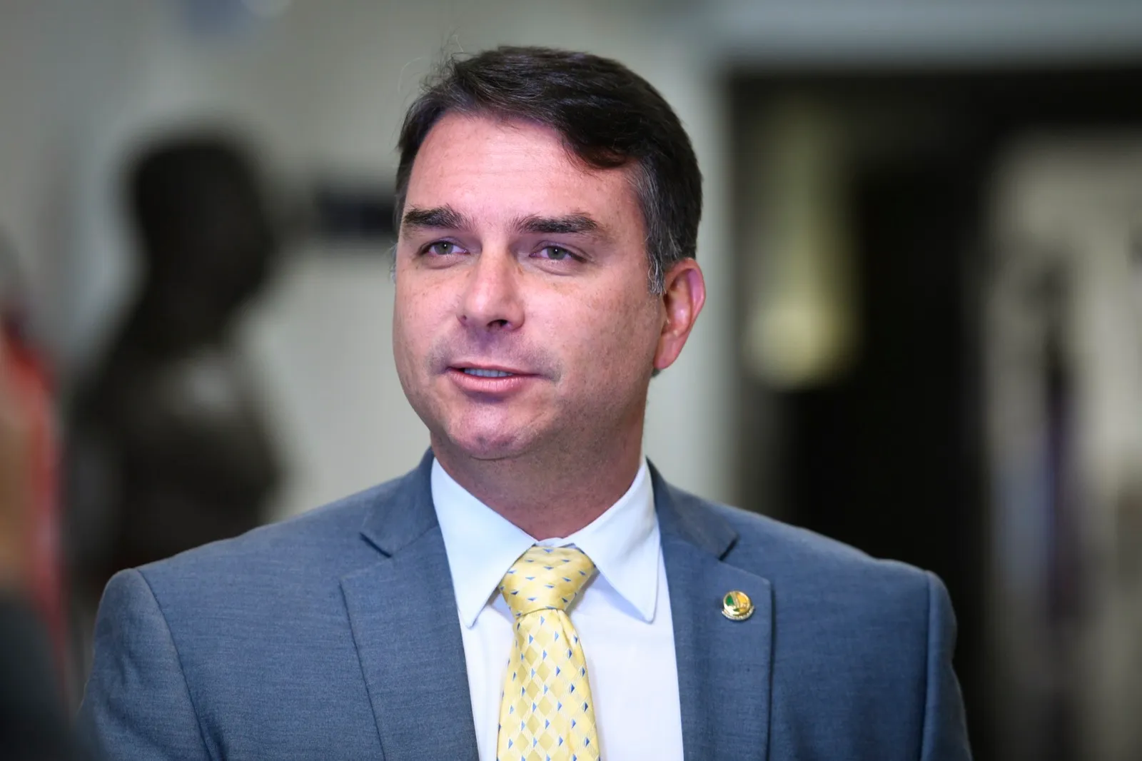 O senador Flávio Bolsonaro, de terno cinza e gravata amarela, fala durante entrevista.