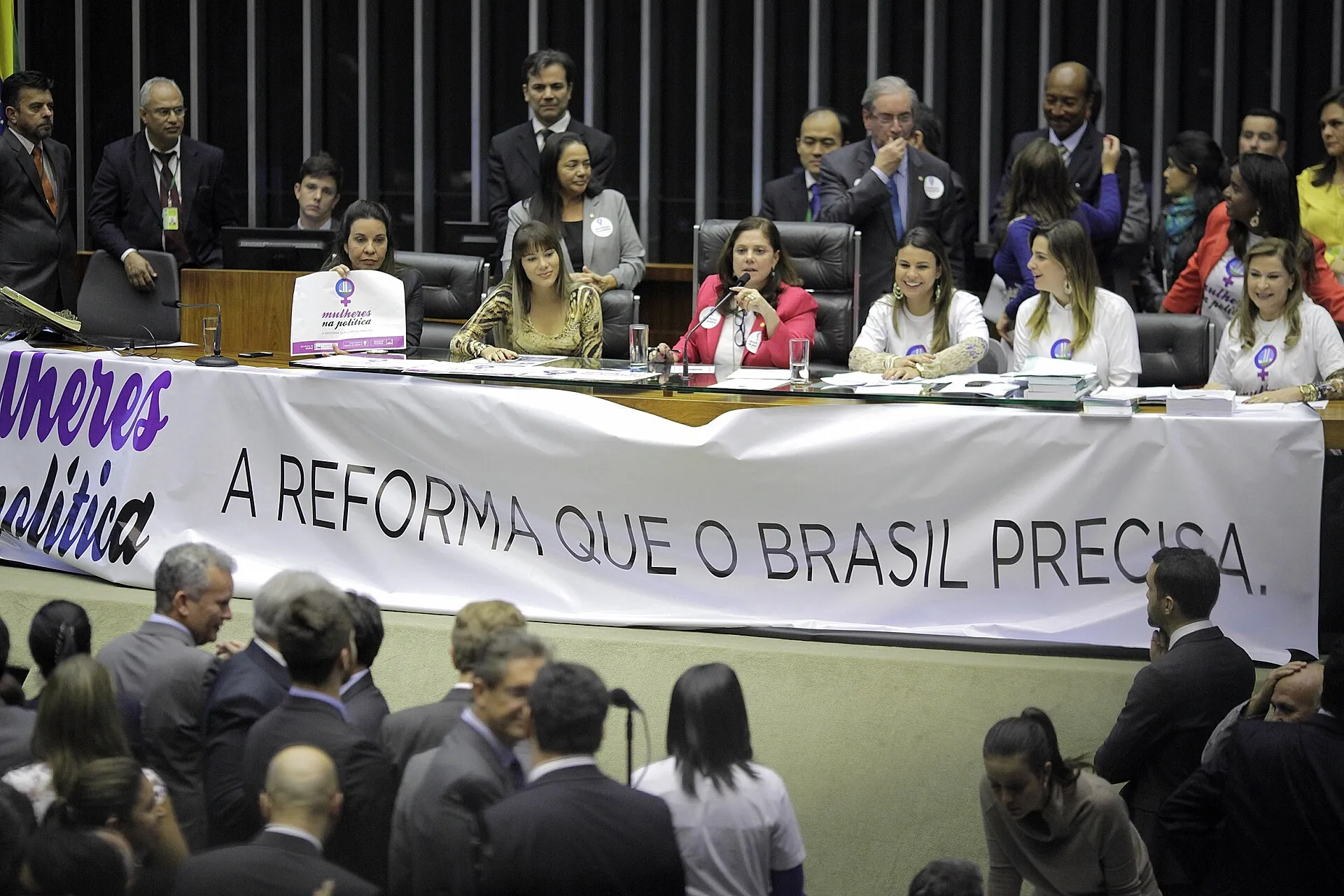 Mesa diretora do Plenário da Câmara dos Deputados com mulheres parlamentares de camiseta branca e uma faixa branca à frente, enquanto deputados conversam em pé no plenário.