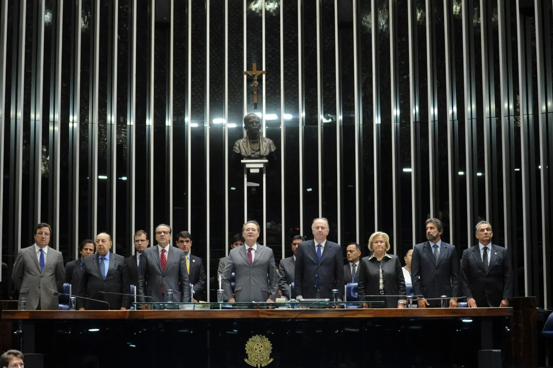 Parlamentares de pé na Mesa do Plenário do Senado, com um busto e uma cruz ao fundo, durante uma sessão solene do Congresso Nacional.