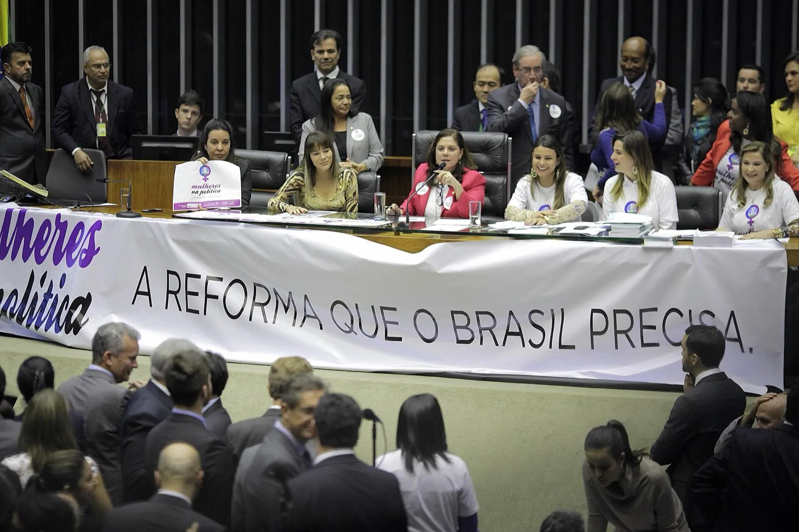 Mesa diretora do plenário da Câmara dos Deputados, com parlamentares sentadas atrás de uma faixa branca com a frase “A reforma que o Brasil precisa”, e deputados em pé no plenário.