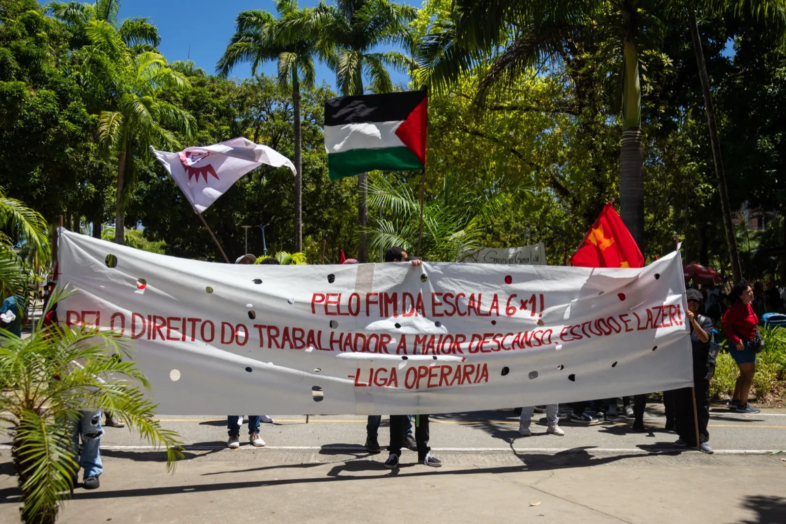 Manifestantes seguram uma faixa branca com os dizeres 'Pelo fim da escala 6x1! Pelo direito do trabalhador a maior descanso, estudo e lazer!', com bandeiras ao fundo.