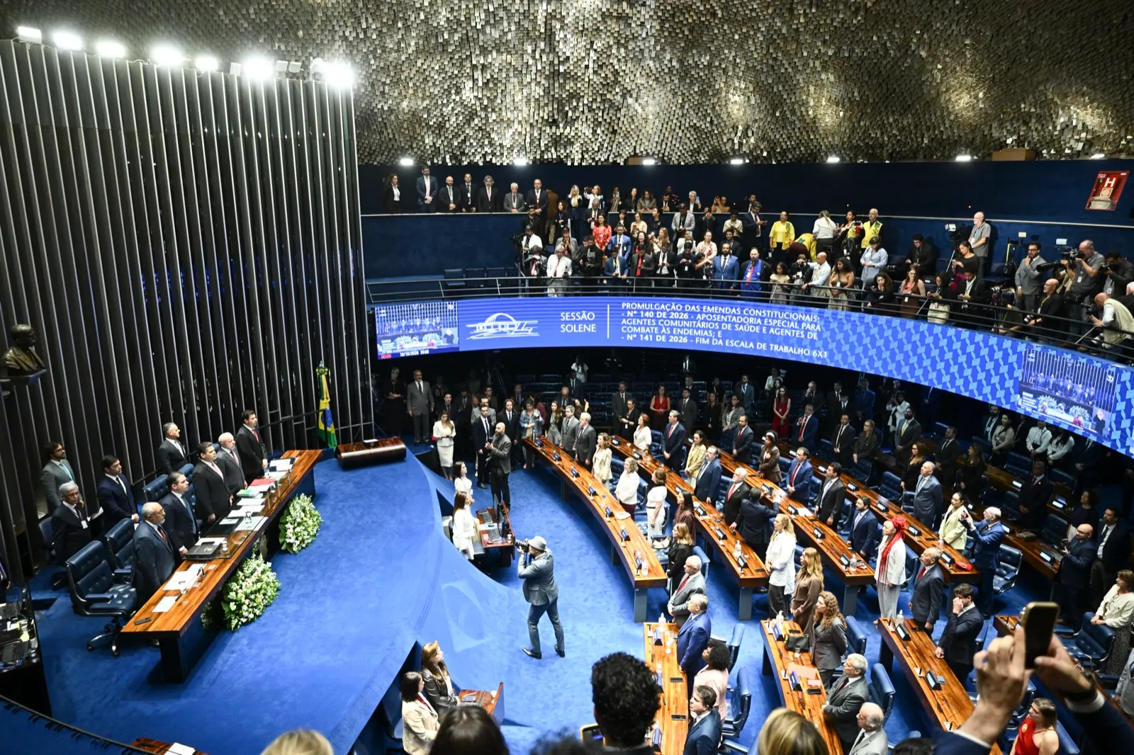 Vista do alto do Plenário do Senado Federal cheio, com parlamentares e convidados nas bancadas e um telão azul ao fundo, durante sessão solene.