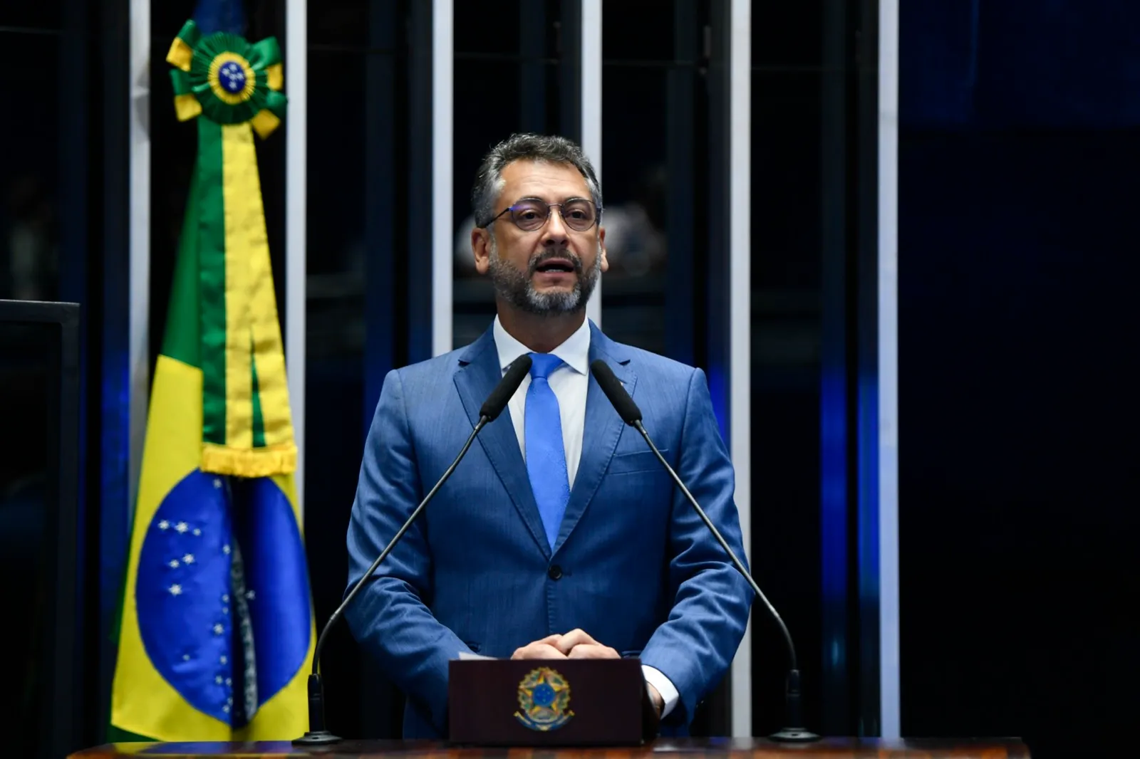 Homem de terno azul discursa na tribuna do Plenário do Senado, ao lado da bandeira do Brasil.