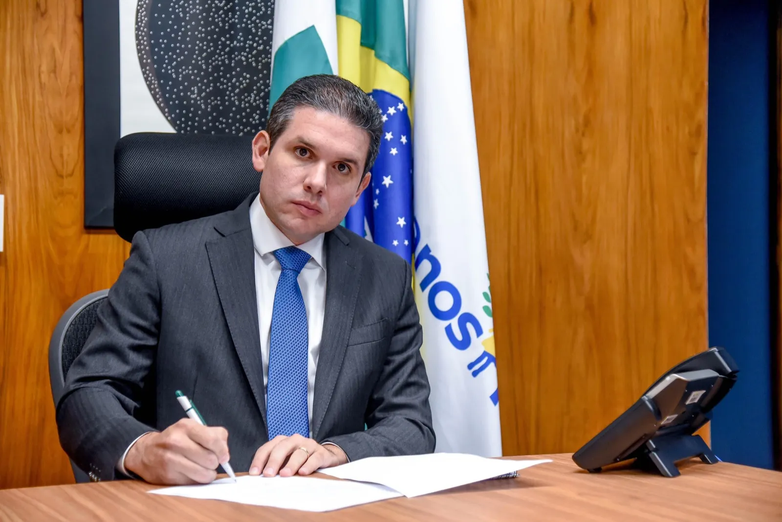 Homem de terno cinza e gravata azul assina papéis em uma mesa, com a bandeira do Brasil ao fundo.