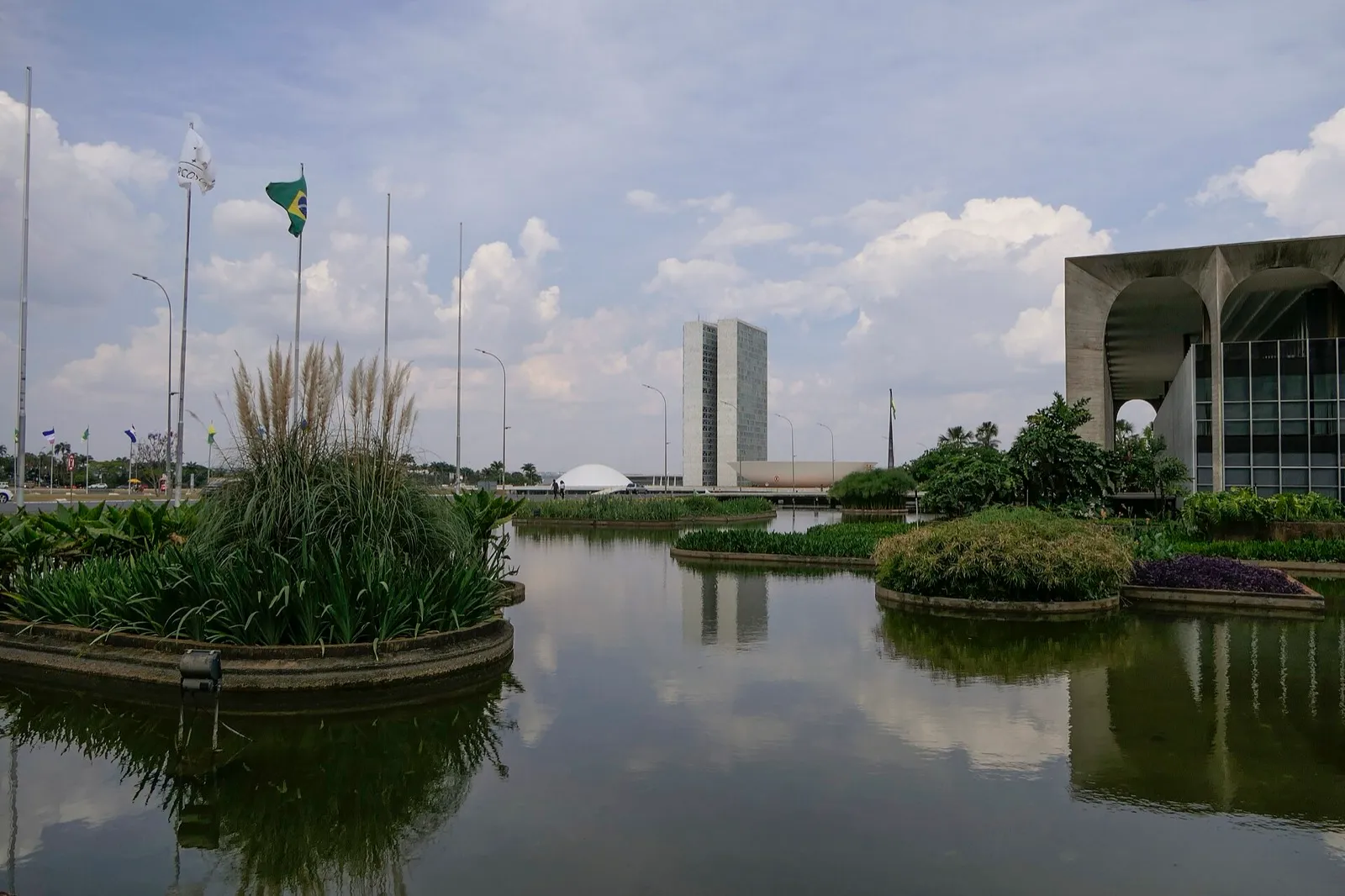 Vista do Congresso Nacional, em Brasília, com as duas torres ao centro e a cúpula branca à esquerda, refletidas em um espelho d'água.