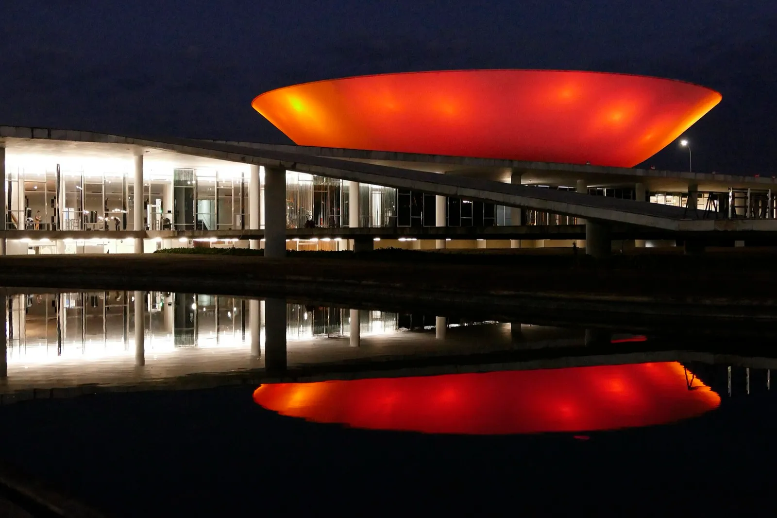 Prédio do Congresso Nacional, em Brasília, iluminado à noite, com uma estrutura curva iluminada em vermelho refletida na água.