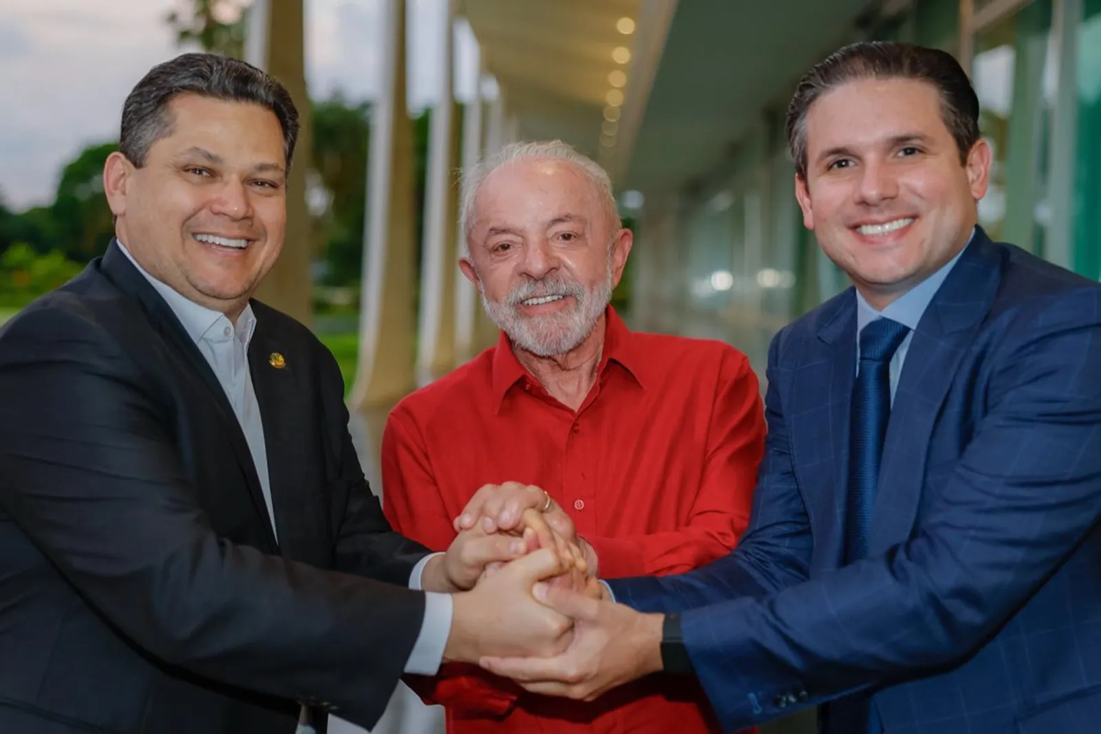 Lula, de camisa vermelha, ao centro, junta as mãos com dois homens de terno no Palácio da Alvorada.