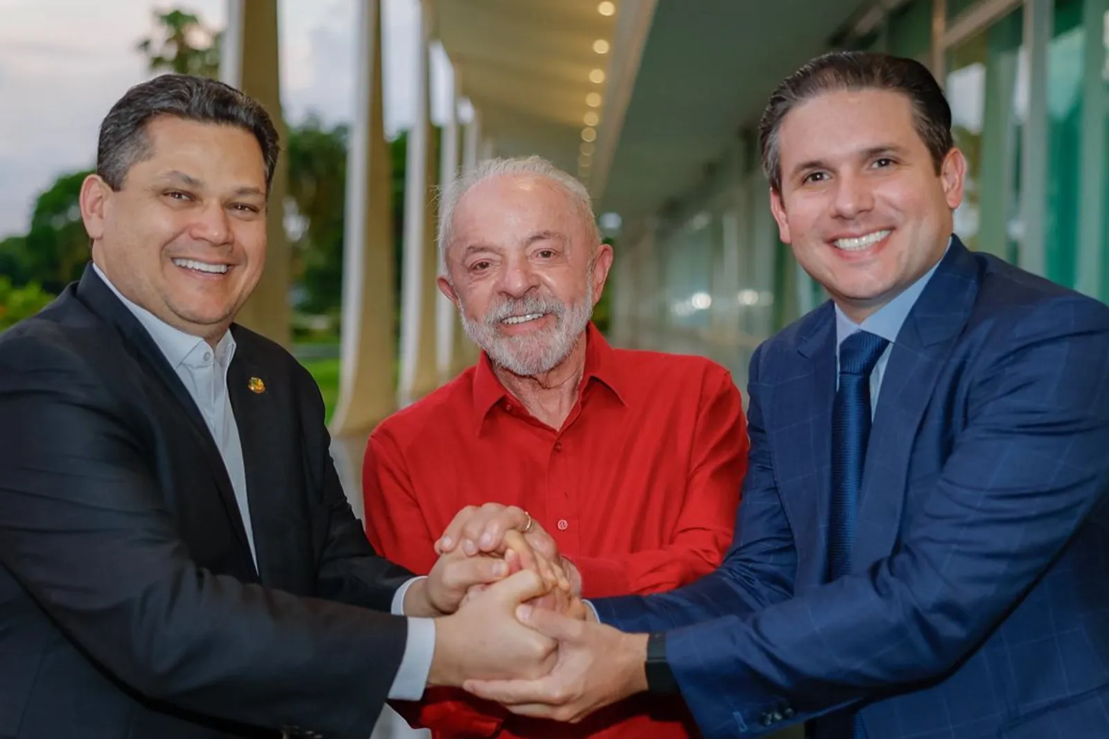 Três homens de mãos dadas em um corredor com colunas: Davi Alcolumbre à esquerda, Lula de camisa vermelha no centro e Hugo Motta à direita.