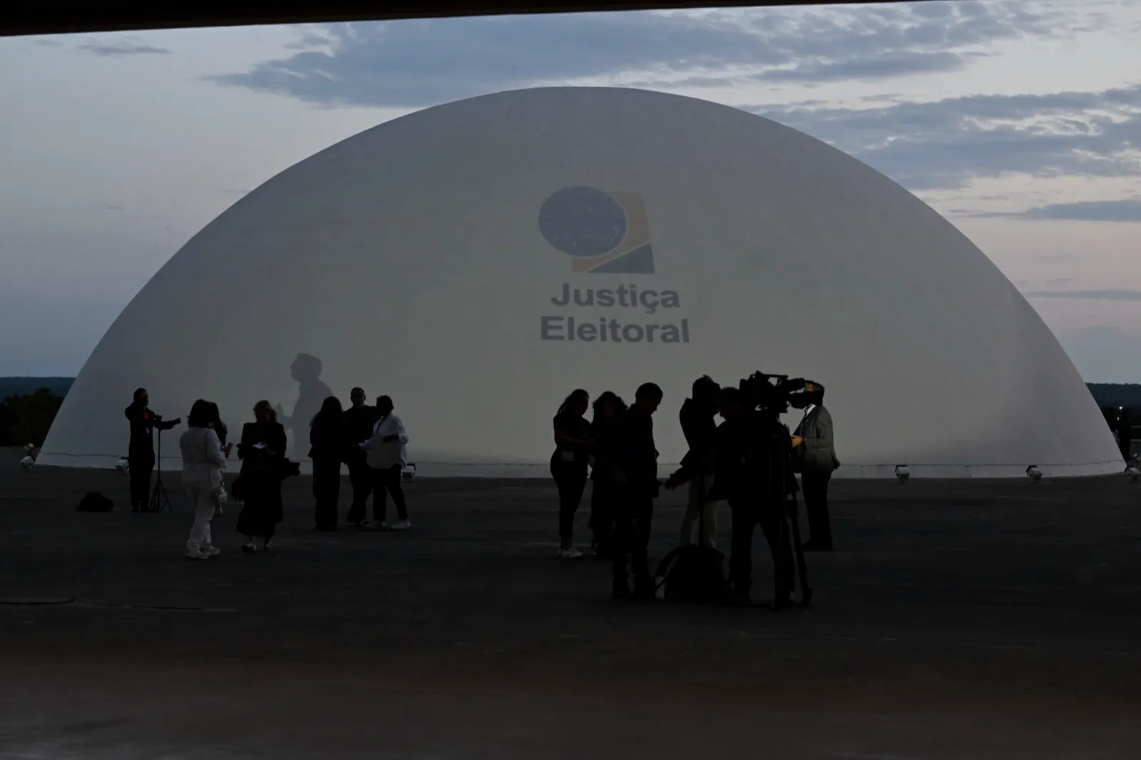 Cúpula branca do Senado Federal, em Brasília, com o logotipo da Justiça Eleitoral projetado, diante de jornalistas e câmeras ao anoitecer.