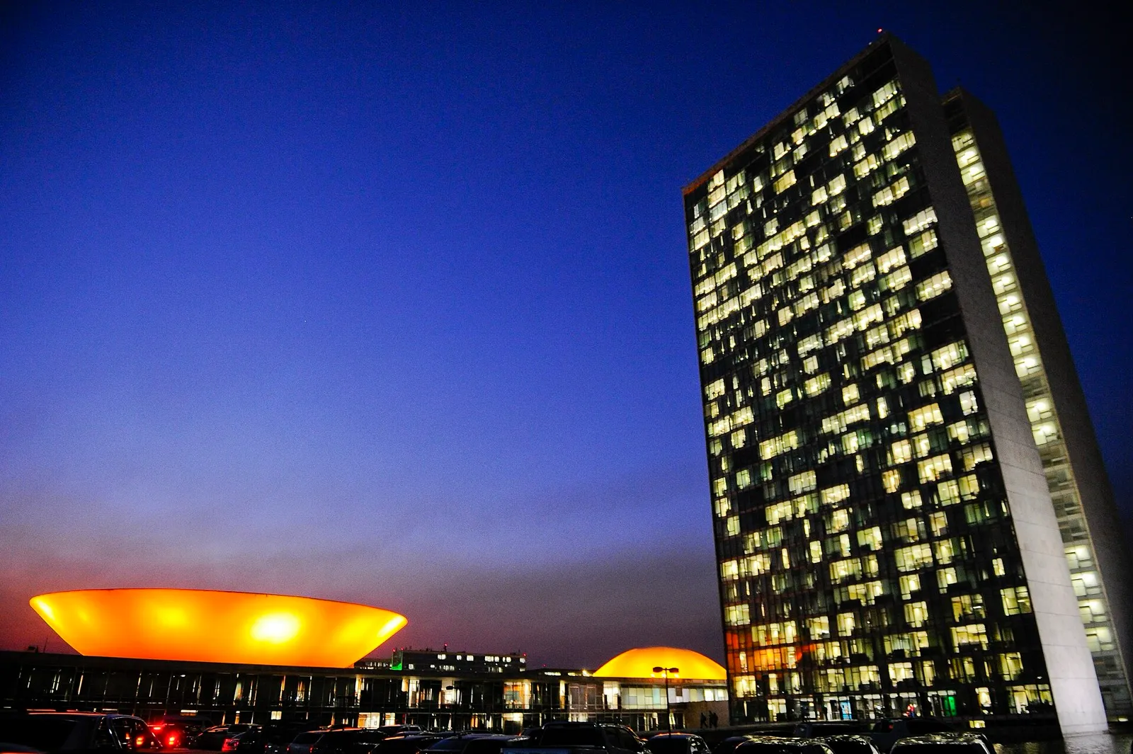 Vista noturna do Congresso Nacional, em Brasília: à esquerda, a cúpula laranja iluminada; à direita, a torre de gabinetes com janelas acesas.
