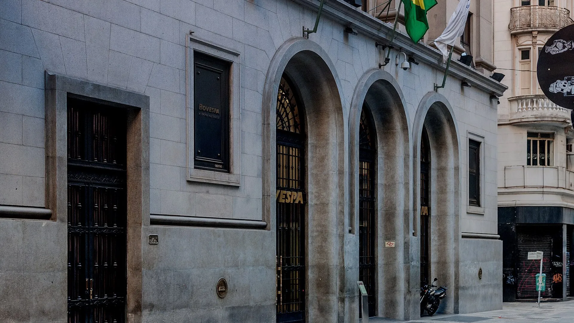 Fachada de pedra da sede da B3 com três arcos de entrada, portões de ferro e a placa Bovespa, em São Paulo.