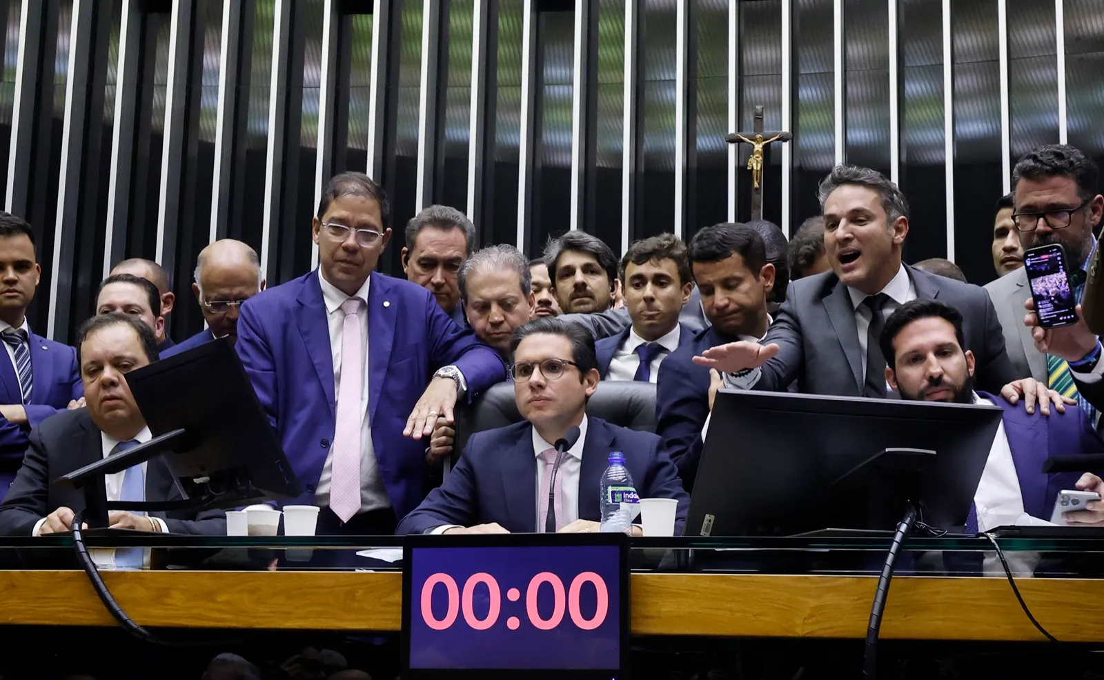 Hugo Motta sentado à mesa do plenário da Câmara, com deputados em pé atrás dele; o painel de tempo marca 00:00.