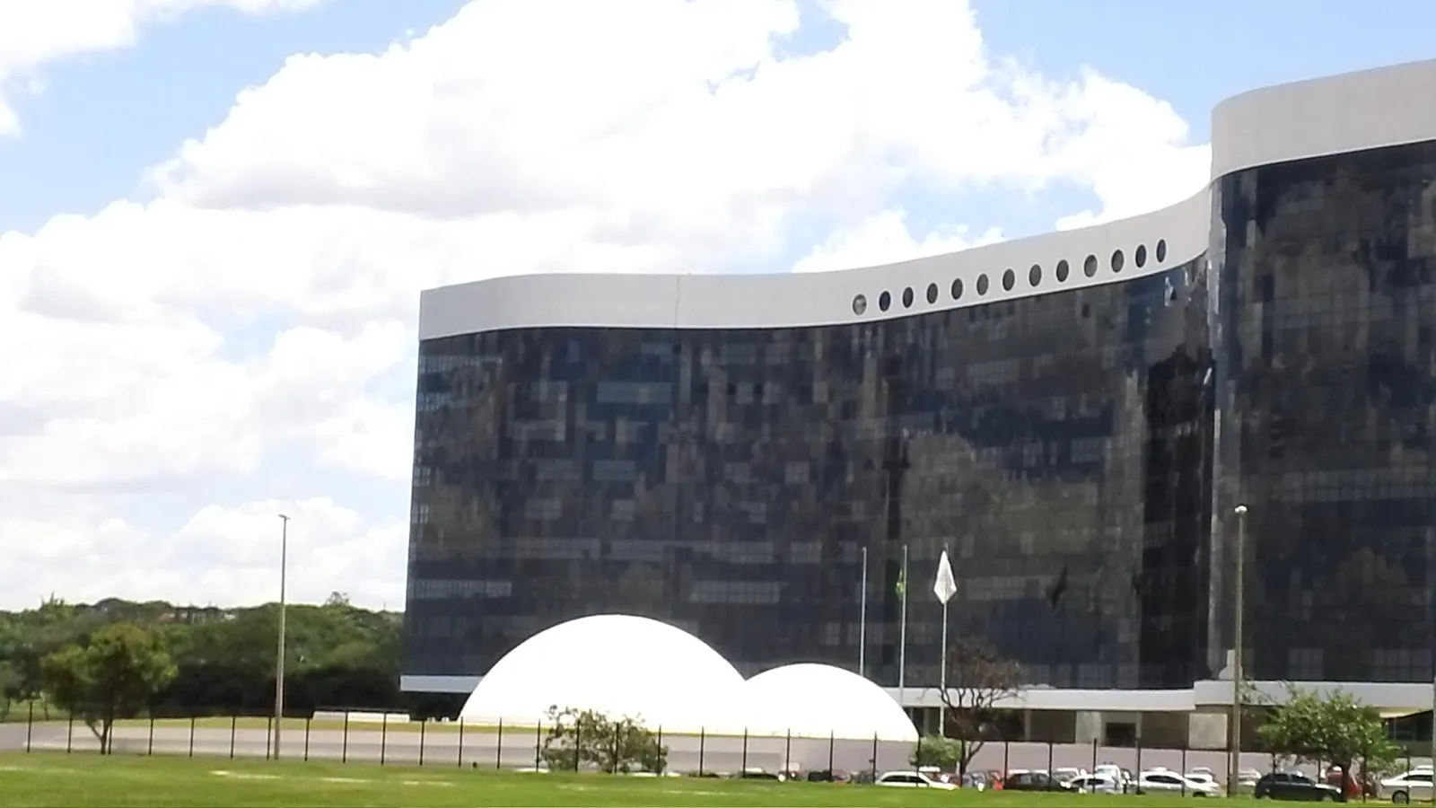 Fachada curva de vidro escuro do edifício do Tribunal Superior Eleitoral, em Brasília, com duas cúpulas brancas na frente e céu com nuvens.