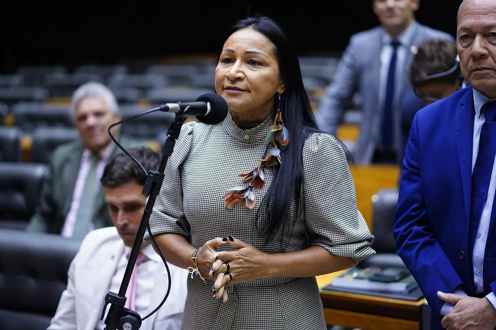 Deputada fala ao microfone no Plenário da Câmara dos Deputados, com cadeiras vazias ao fundo e um homem de terno azul ao lado.