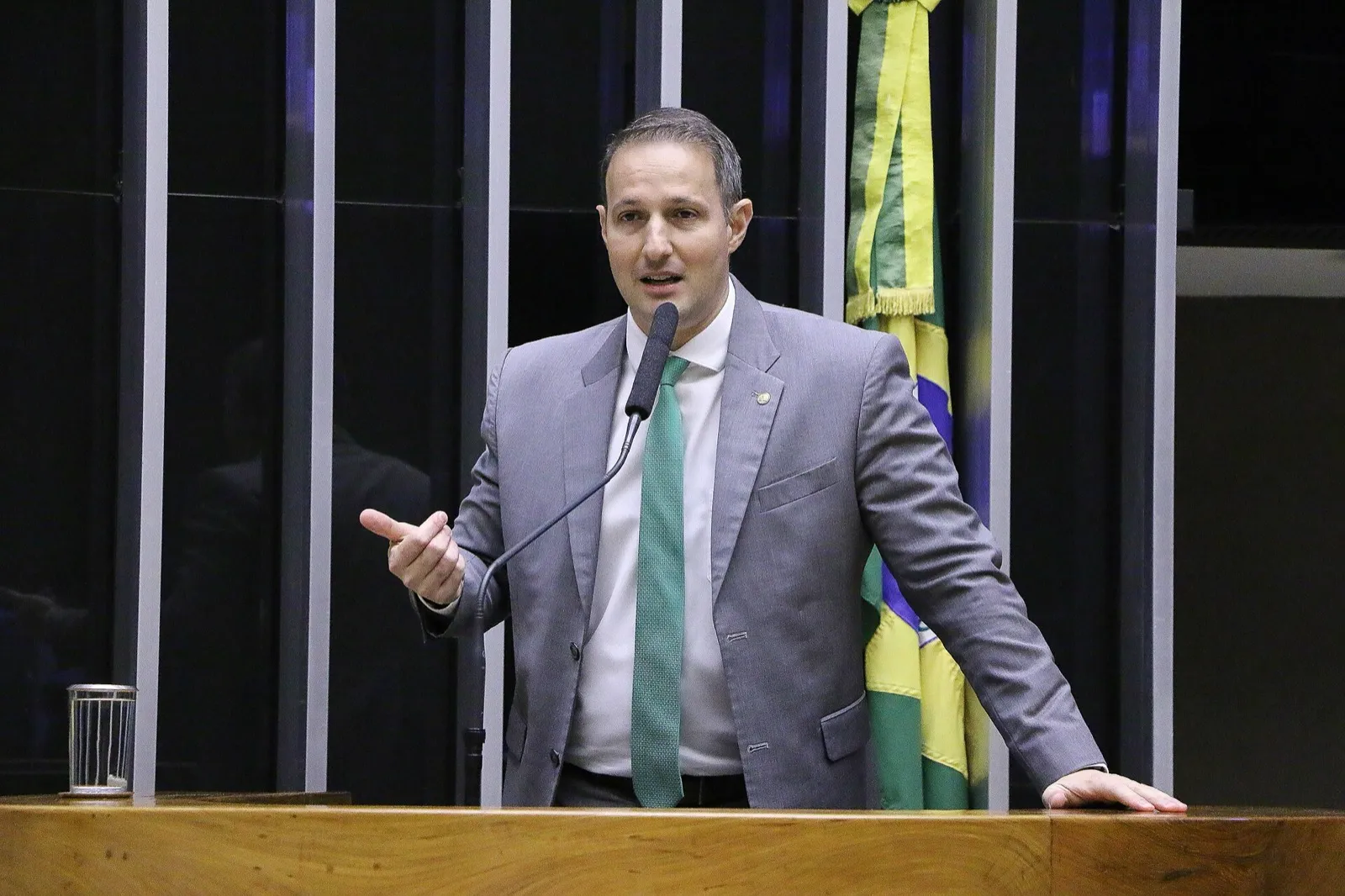 O deputado Guilherme Derrite, de terno cinza e gravata verde, discursa na tribuna do plenário da Câmara, ao lado da bandeira do Brasil.