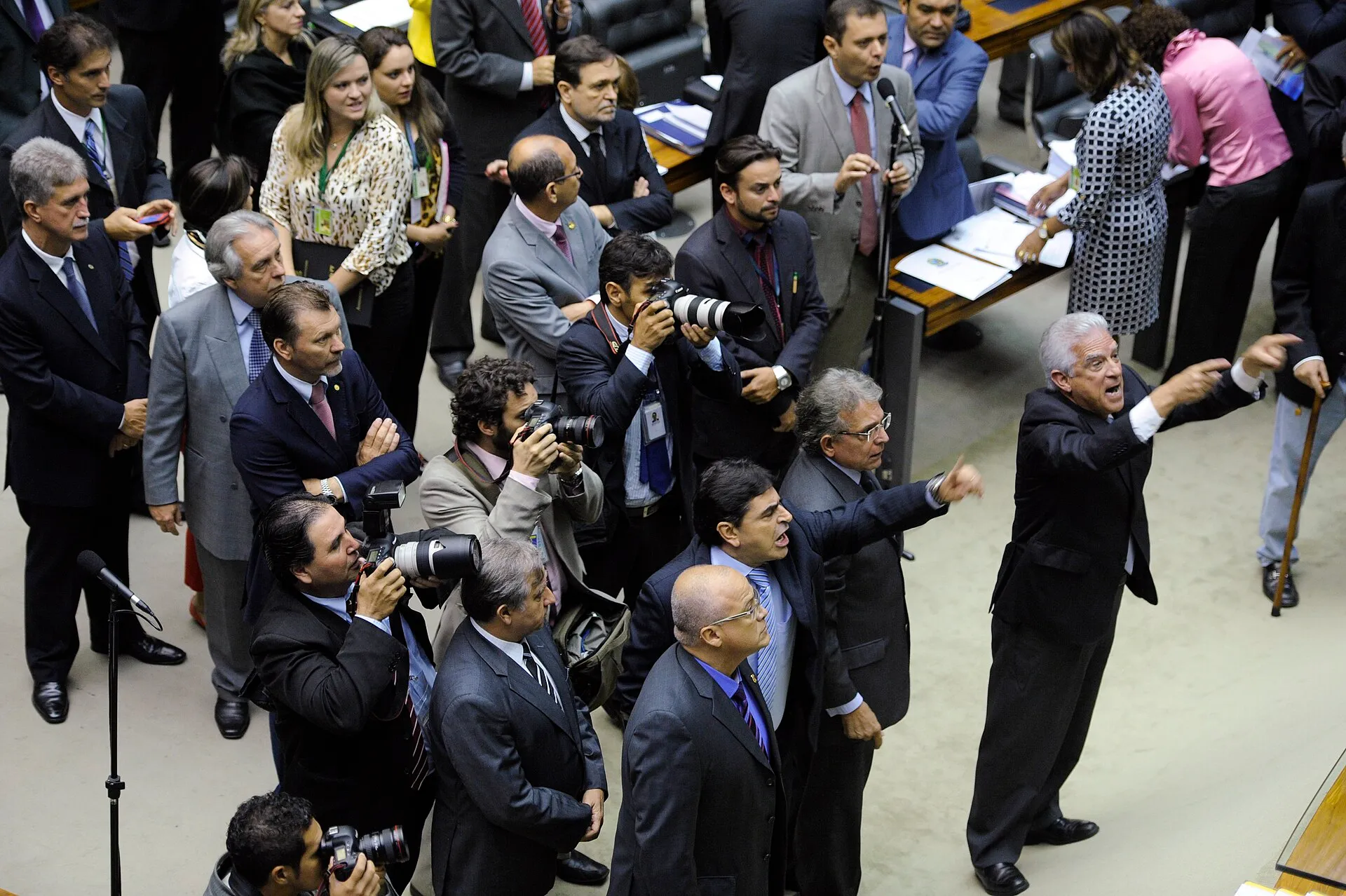 Parlamentares em pé no Plenário, alguns gesticulando, cercados por fotógrafos e assessores, visto do alto.