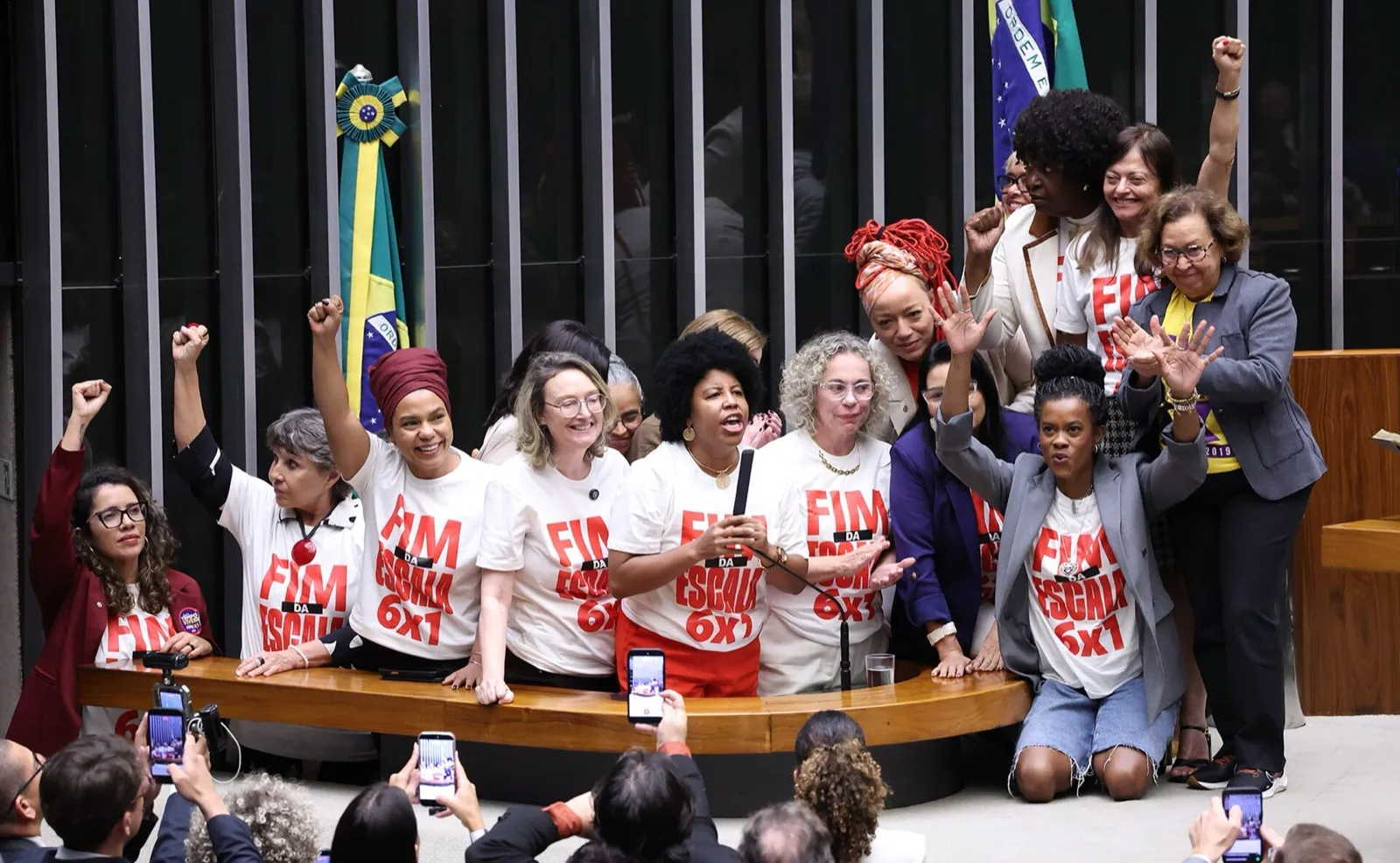 Deputadas da base governista, de camisetas brancas com a frase “Fim da escala 6x1”, comemoram com braços erguidos junto à mesa do Plenário da Câmara.