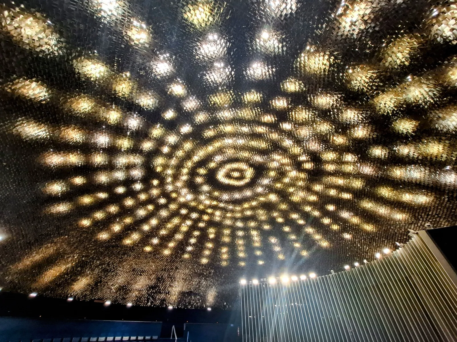 Teto do Plenário do Senado Federal, coberto por peças metálicas douradas iluminadas por dezenas de luzes, com o painel de lâminas verticais ao fundo.