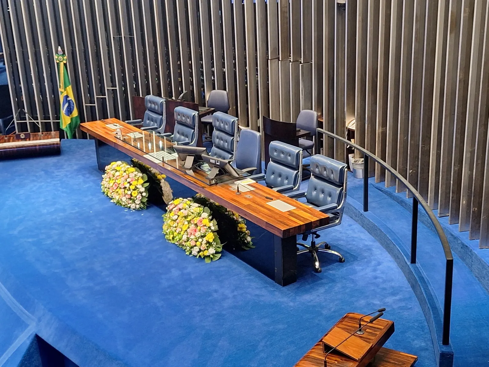 Mesa diretora vazia do Plenário do Senado Federal, com cadeiras azuis, arranjos de flores, a bandeira do Brasil e a tribuna em primeiro plano.