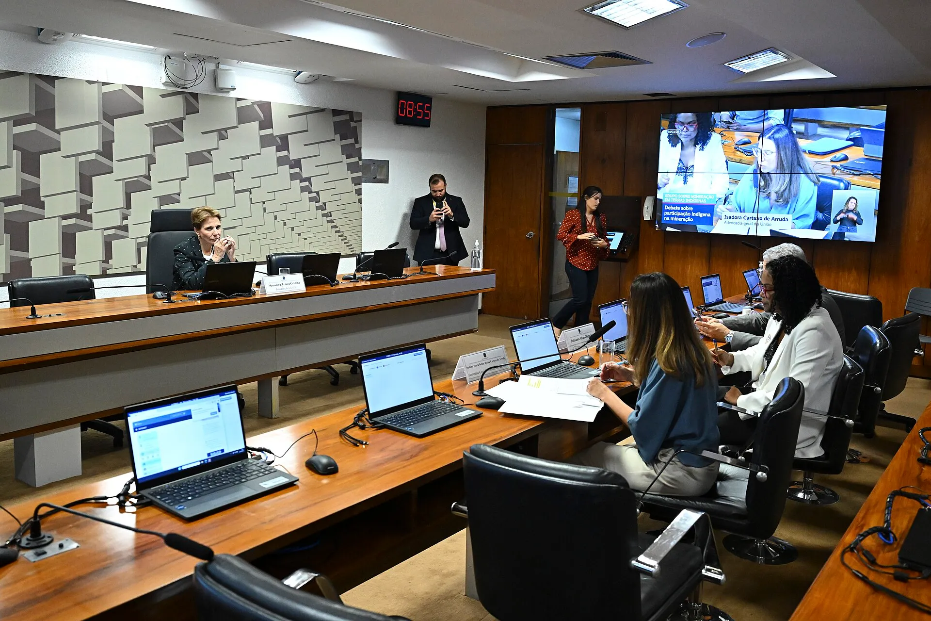 Senadora Tereza Cristina sentada à mesa de uma sala de audiência do Senado, com um telão ao fundo que mostra o nome do debate sobre mineração em terras indígenas.