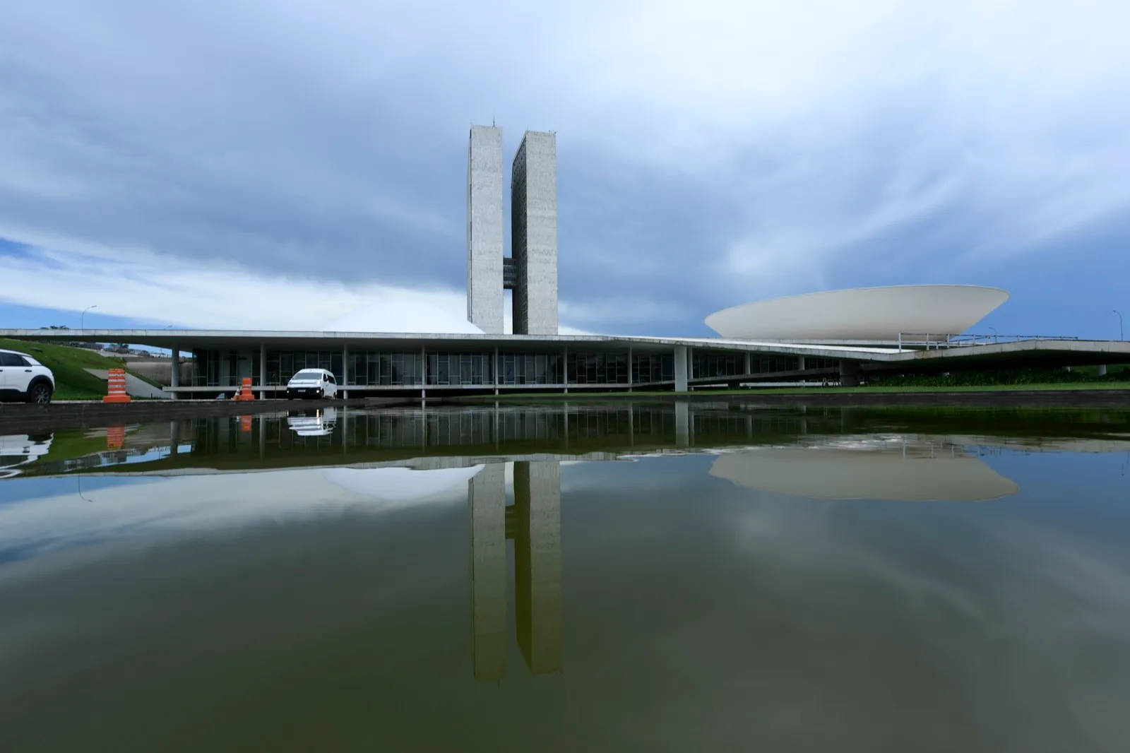 Palácio do Congresso Nacional, com as duas torres e as cúpulas refletidas no espelho d'água, sob céu nublado.