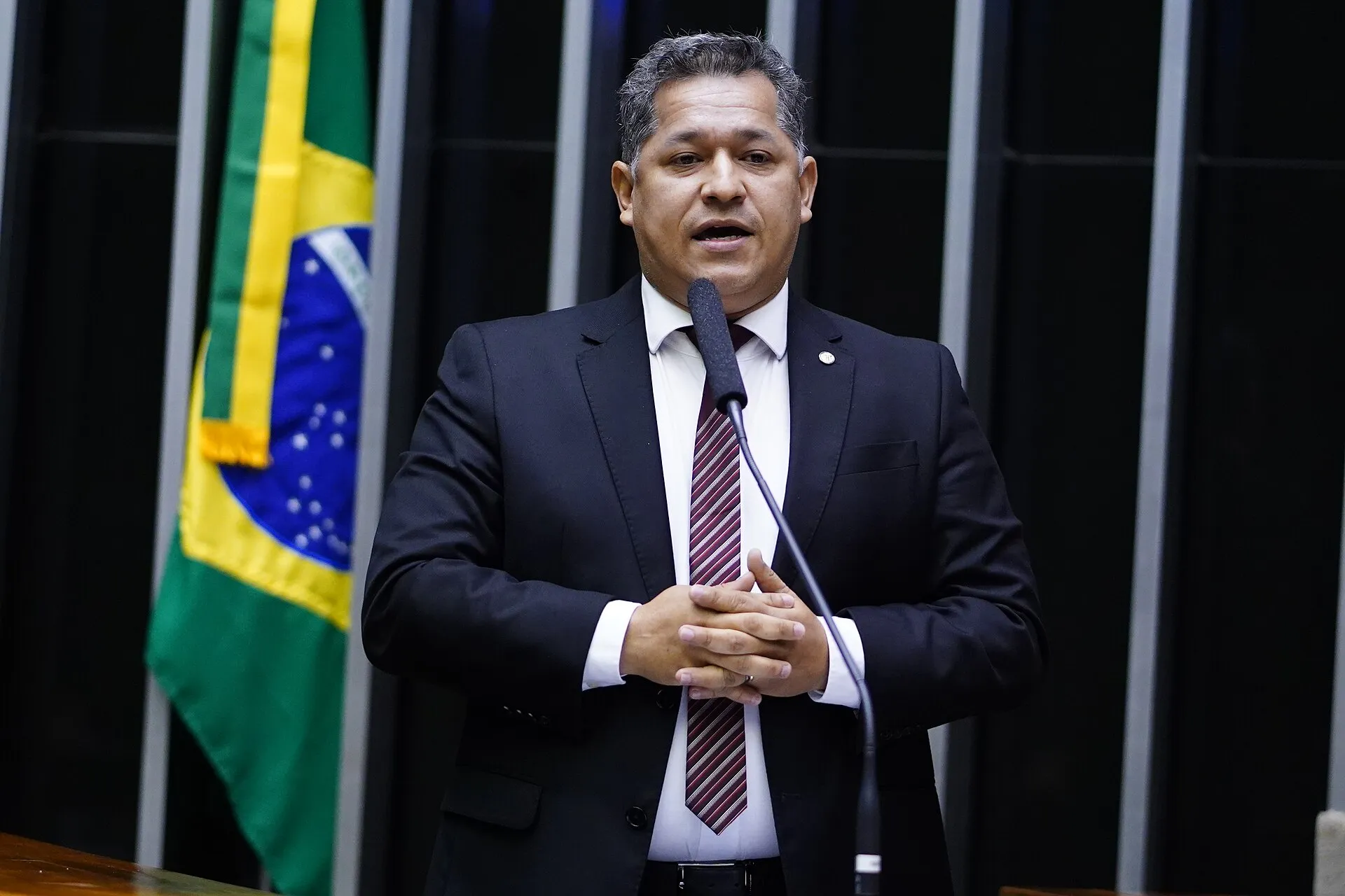 Deputado de terno escuro fala ao microfone na tribuna do Plenário da Câmara dos Deputados, com a bandeira do Brasil ao fundo.