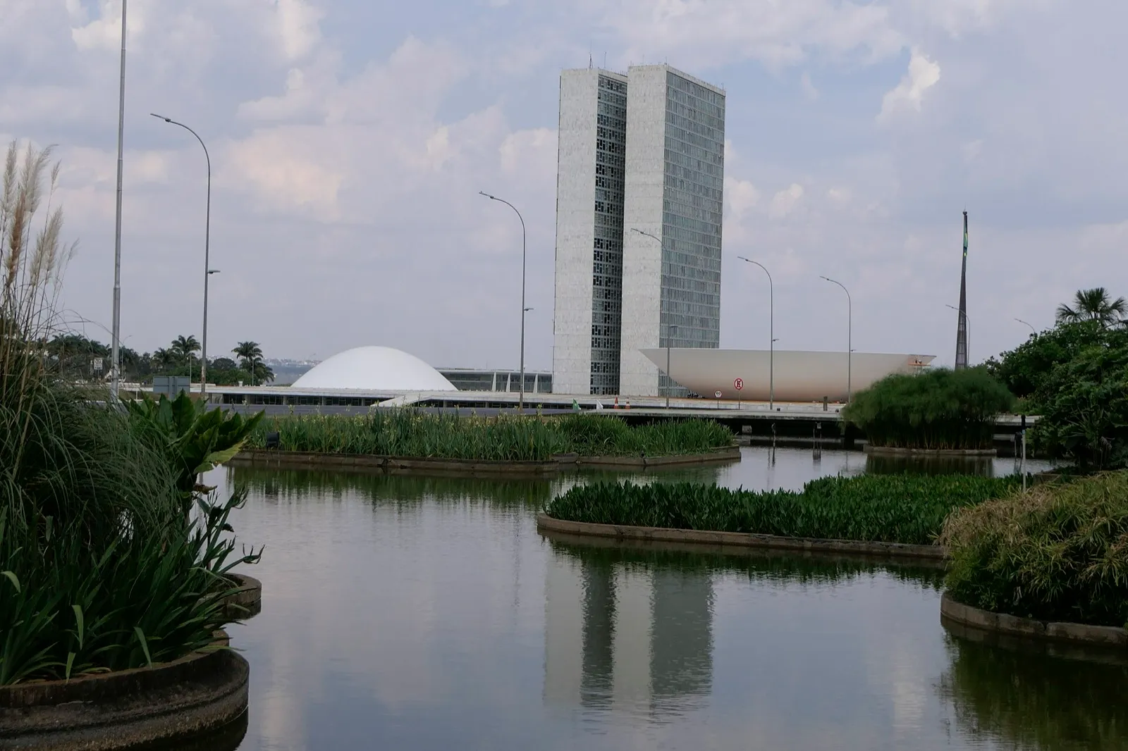 Fachada do Congresso Nacional, em Brasília, com a cúpula branca e as duas torres ao fundo, vista de um espelho d'água com vegetação.