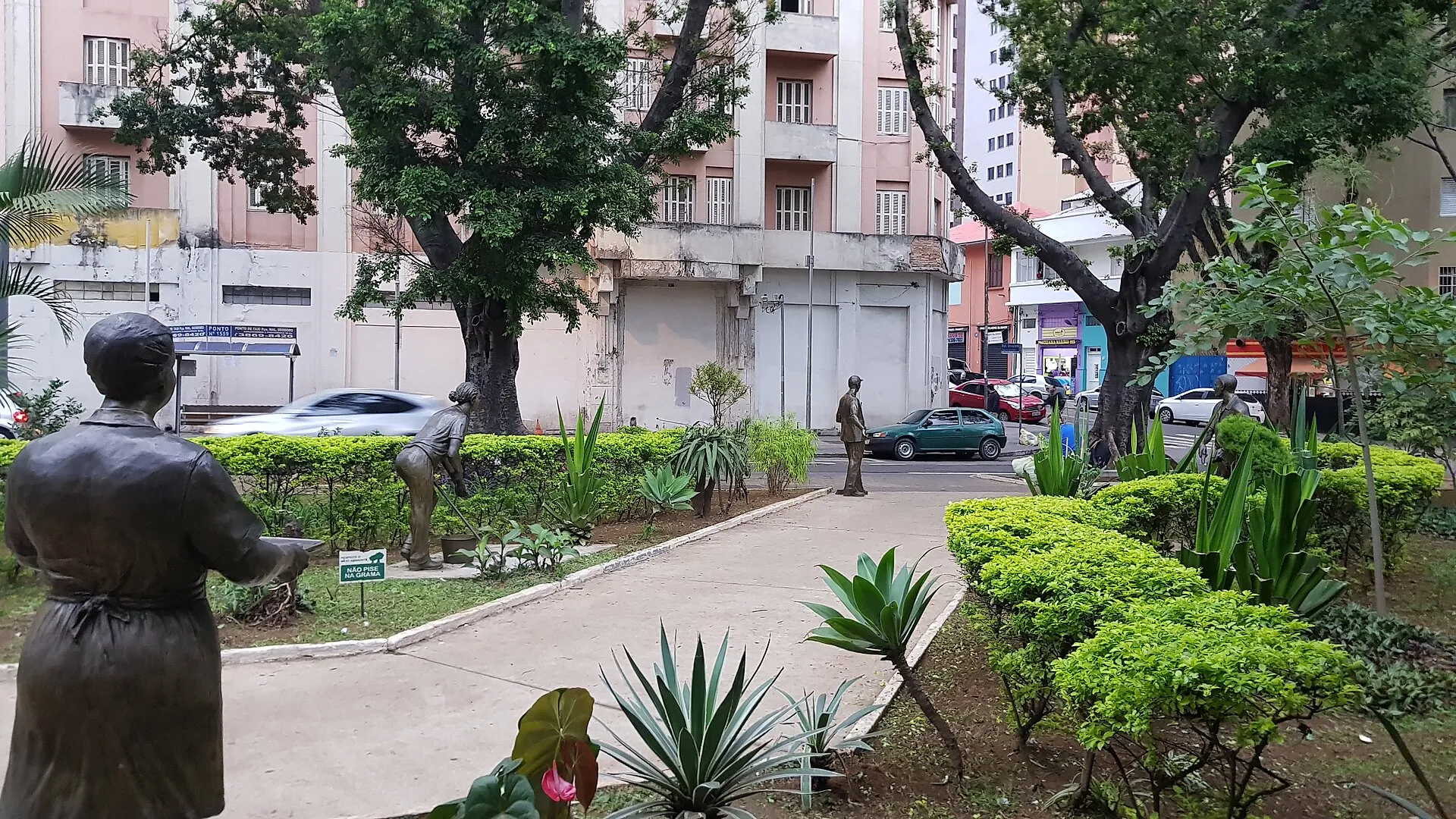 Estátuas de bronze de trabalhadores da limpeza urbana, com um gari curvado sobre a calçada, em uma praça de São Paulo, com prédios, árvores e carros ao fundo.