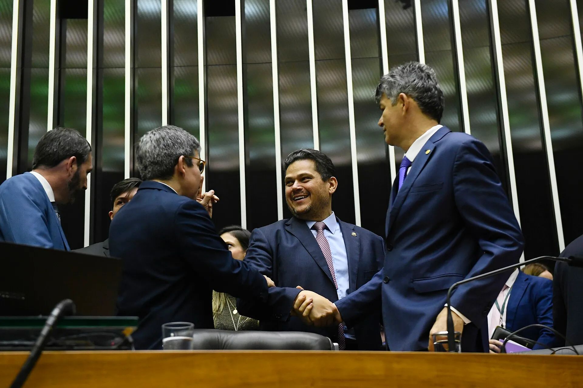 Homens de terno escuro cumprimentam-se na mesa do plenário da Câmara dos Deputados, com painéis escuros e brancos ao fundo.