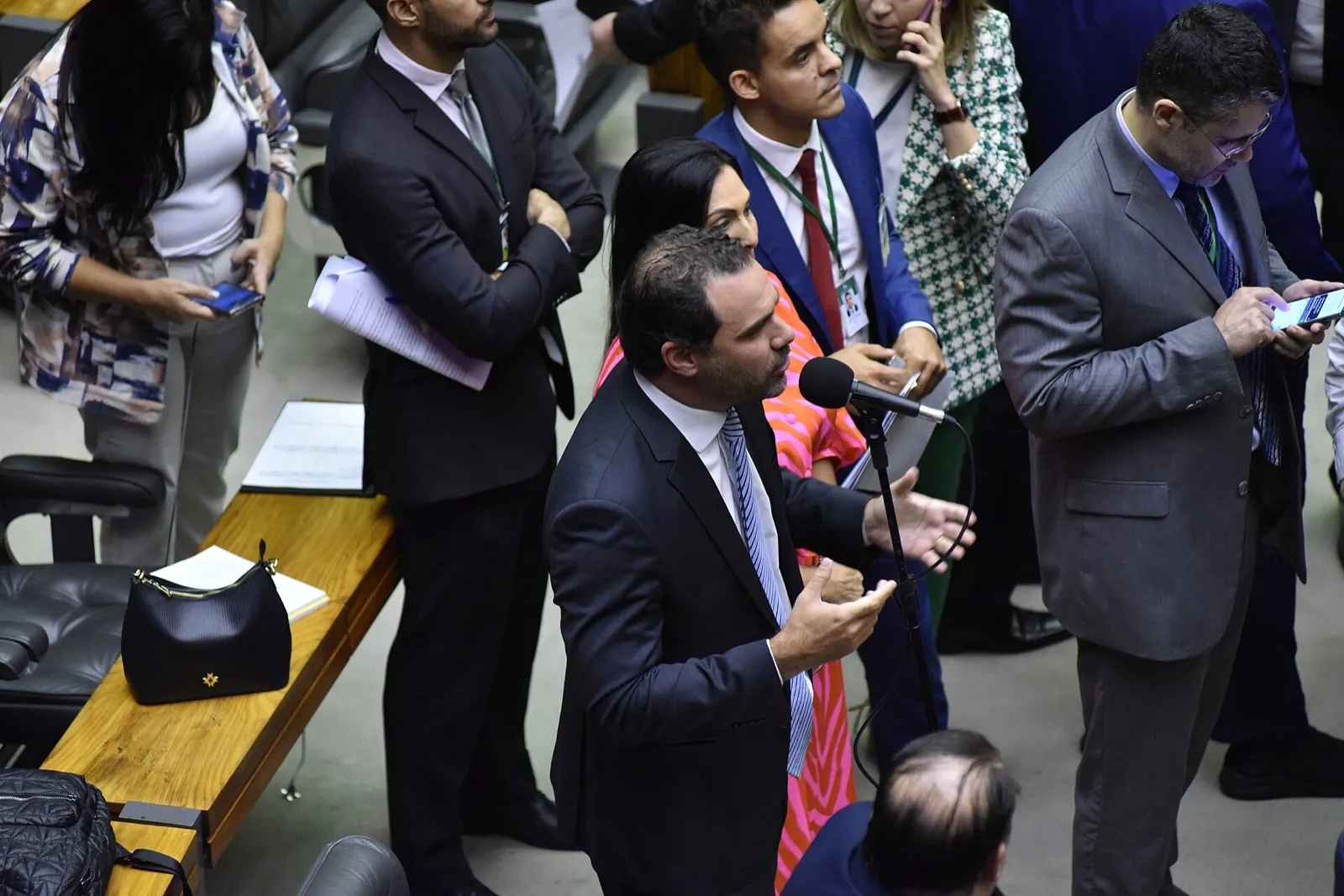 Deputado de terno escuro fala ao microfone no plenário da Câmara dos Deputados, cercado de pessoas em pé.