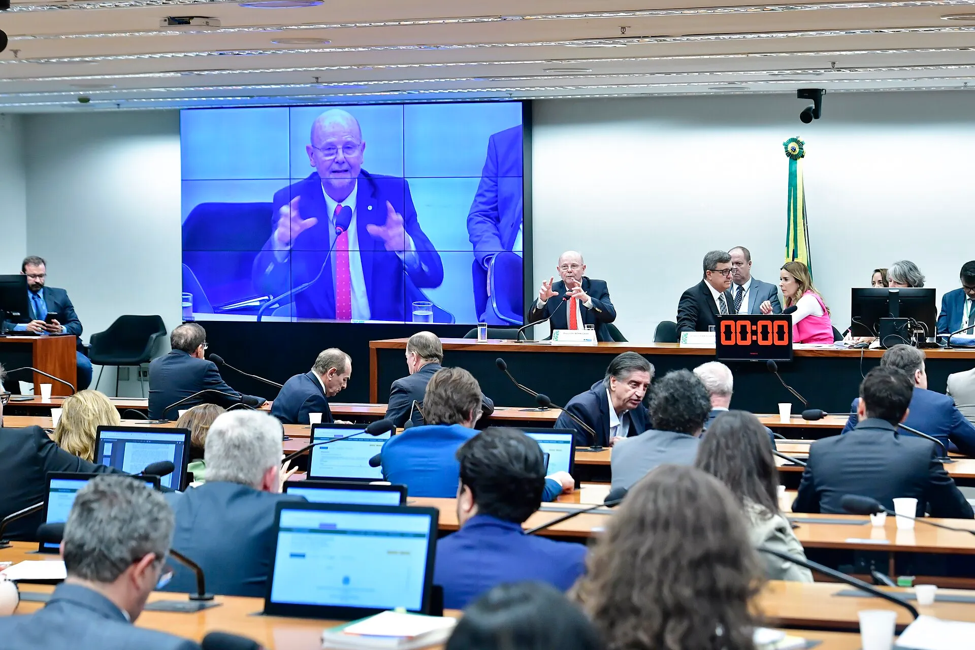 Reunião da Comissão Mista de Orçamento, com a mesa diretora ao fundo, um telão exibindo o orador e parlamentares sentados diante de notebooks.