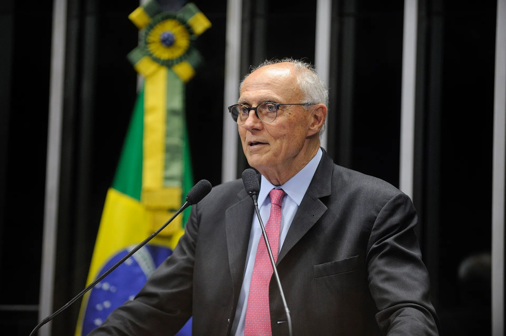 Senador de cabelos grisalhos, óculos e terno escuro fala ao microfone na tribuna do Plenário do Senado, com a bandeira do Brasil ao fundo.