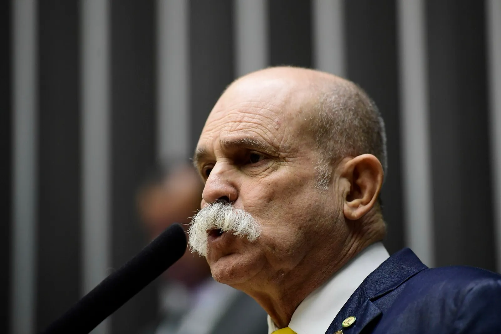 Deputado discursa ao microfone no plenário da Câmara, com painéis escuros listrados ao fundo.