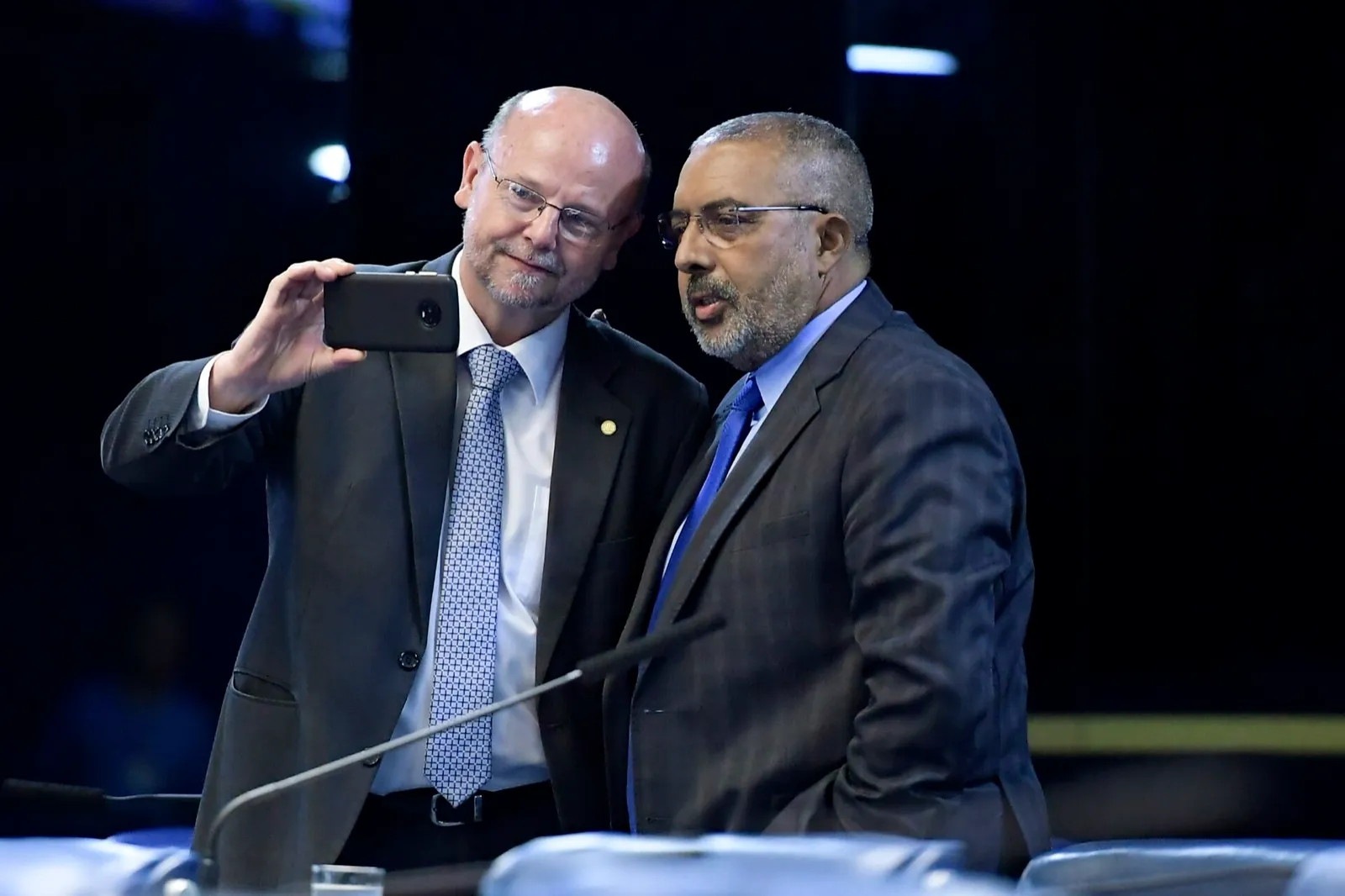 Dois homens de terno no Plenário do Senado. À esquerda, o deputado Bohn Gass tira uma selfie com o celular; à direita, o senador Paulo Paim, de óculos.