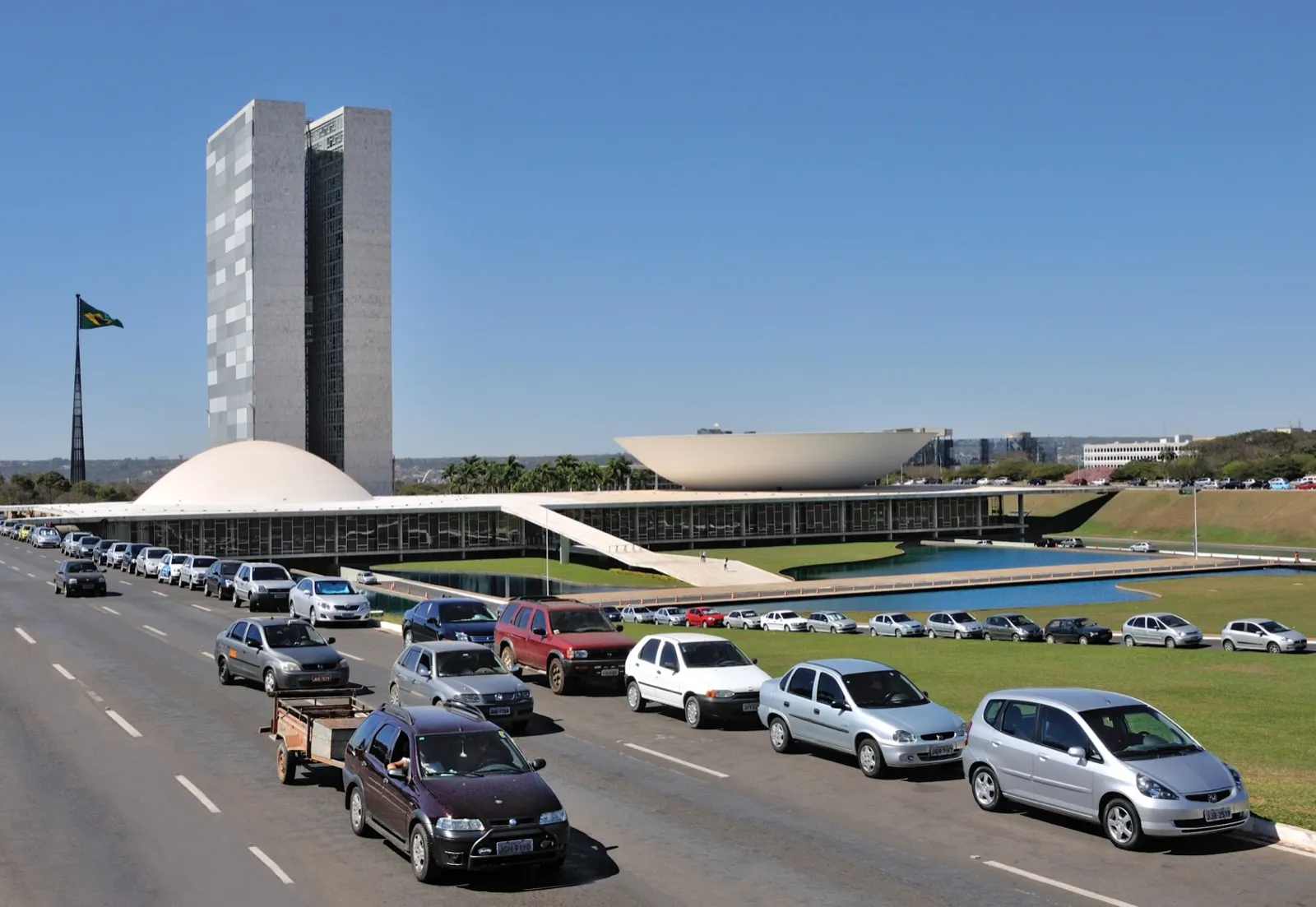 Palácio do Congresso Nacional, em Brasília, com as duas torres e as cúpulas brancas, e uma fila de carros na avenida em primeiro plano.