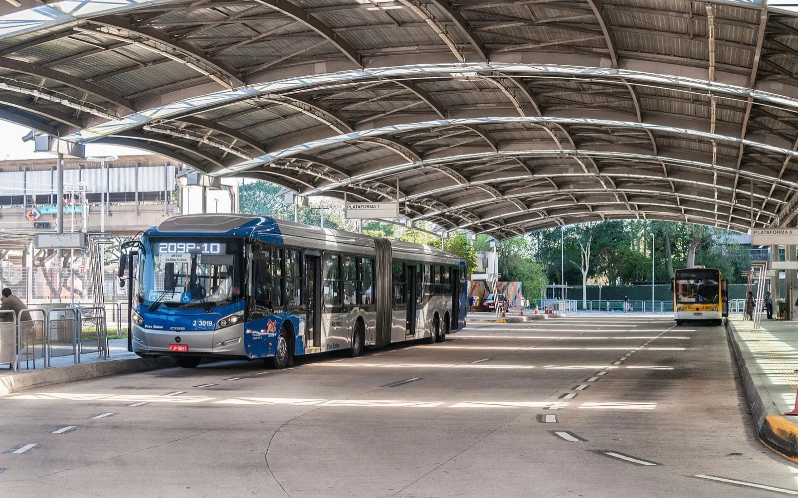 Ônibus articulado azul e prata em um terminal de São Paulo, sob uma cobertura metálica curva, com outro ônibus ao fundo.