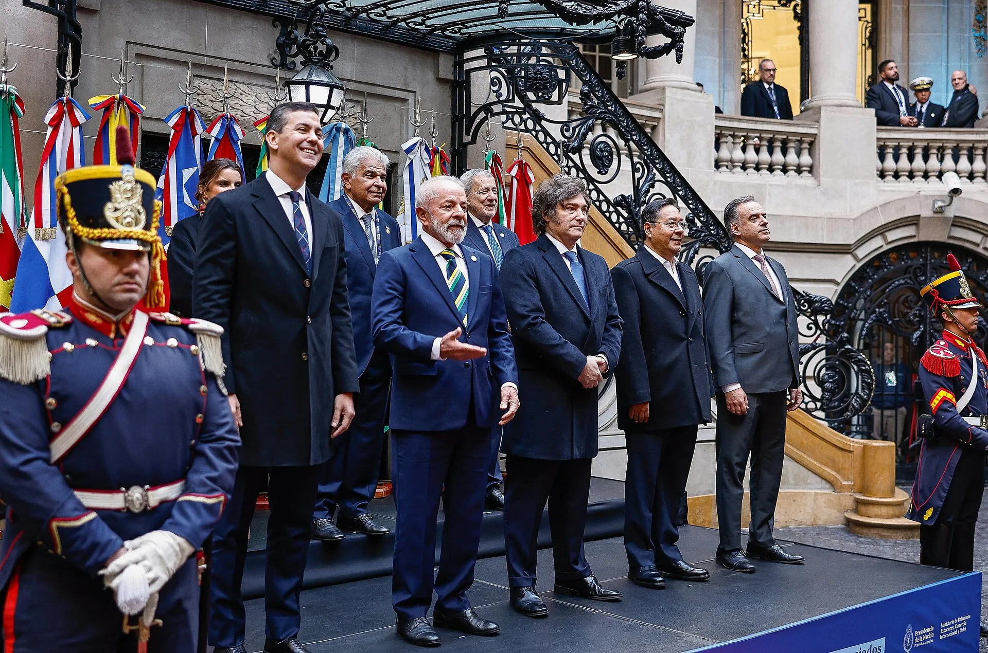 Lula e outros chefes de Estado posam em fila sobre um palco, com bandeiras ao fundo e guardas de uniforme em primeiro plano.