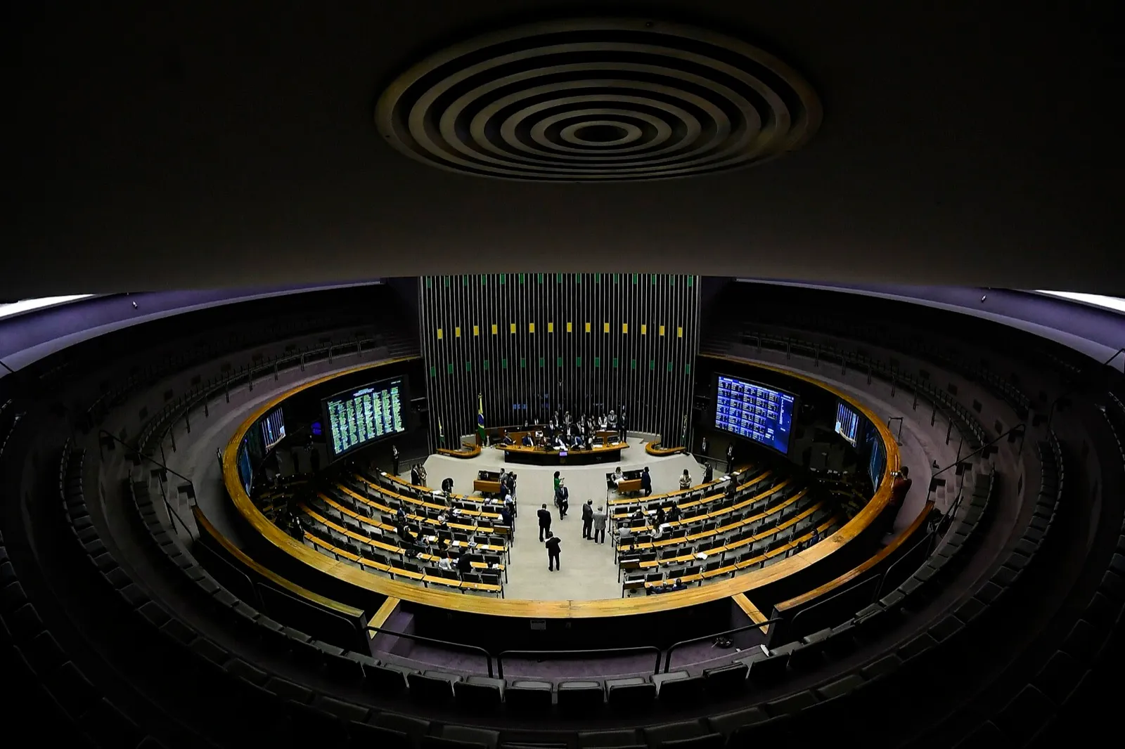 Plenário da Câmara dos Deputados visto do alto, com poucos parlamentares nas bancadas, a mesa diretora ao fundo e painéis de votação acesos nas laterais.